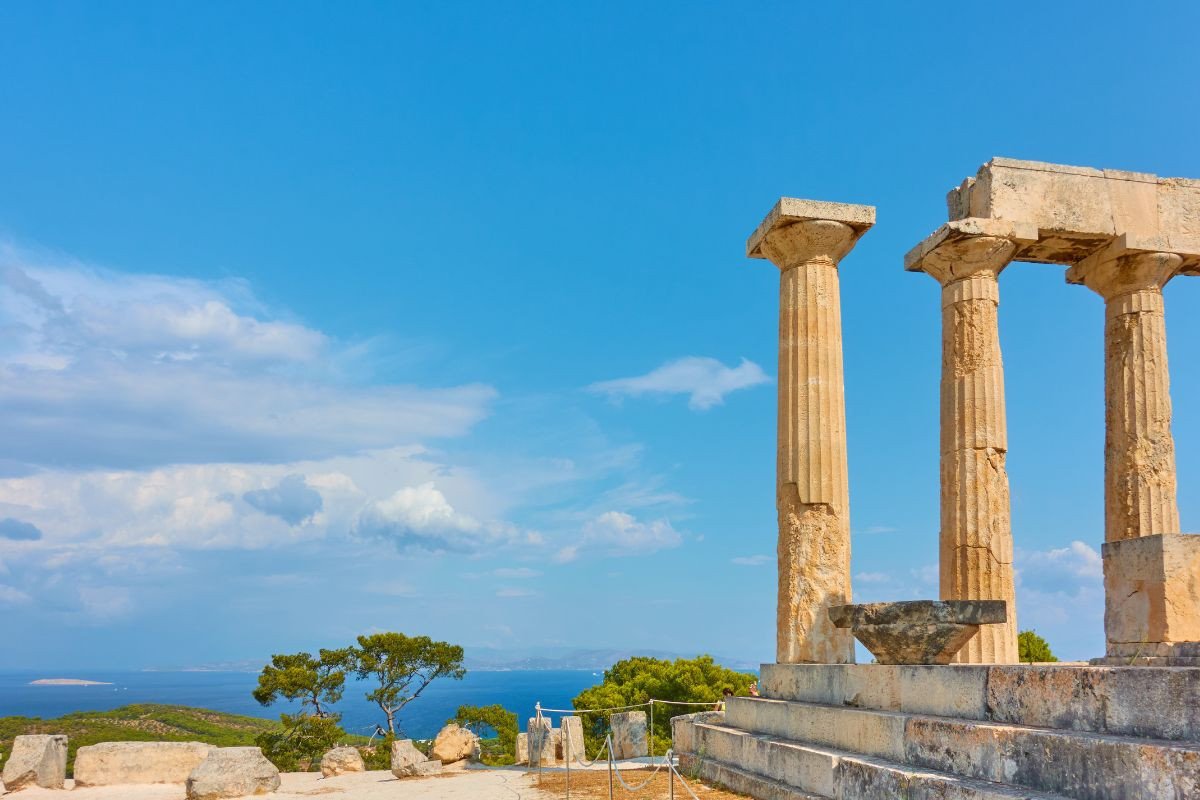 Ancient temple ruins with standing columns on a hilltop overlooking the sea, with a clear blue sky and scattered clouds in the background.